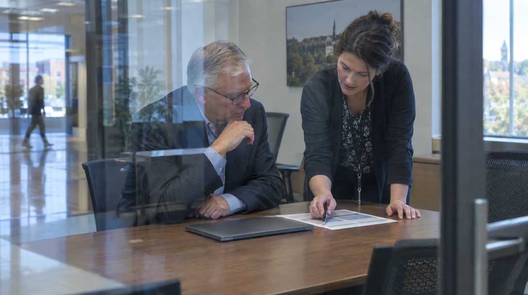 A city clerk and two staff members review printed meeting packets and a contract binder at a conference table in a city hall office.